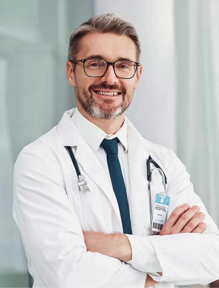 A male doctor with glasses and a beard, wearing a white coat and stethoscope, stands with arms crossed, smiling. He has a hospital ID badge and is indoors in a bright, modern setting.