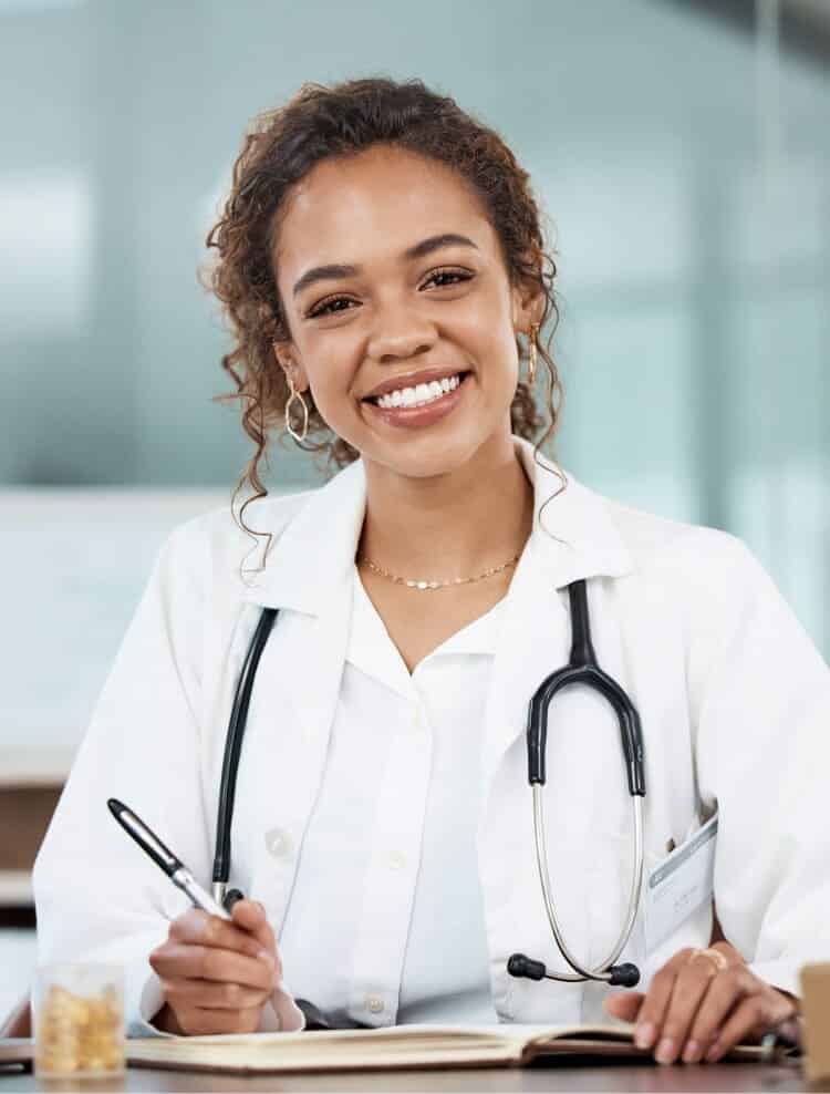A smiling doctor wearing a white coat and stethoscope sits at a desk, holding a pen and writing in a notebook.