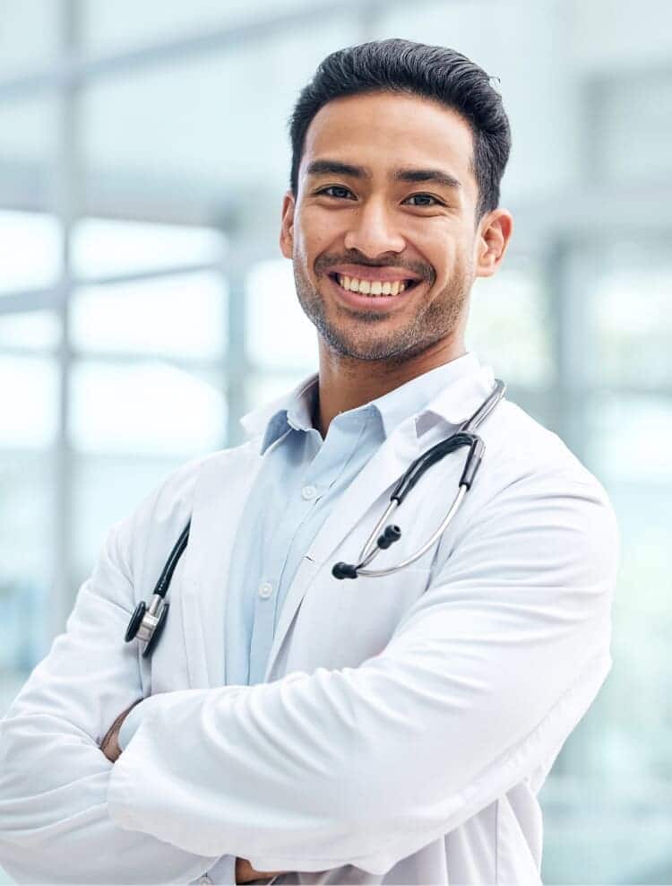 A young male doctor with dark hair smiles confidently, wearing a white lab coat and a stethoscope around his neck. He stands with arms crossed in a bright, modern medical facility.