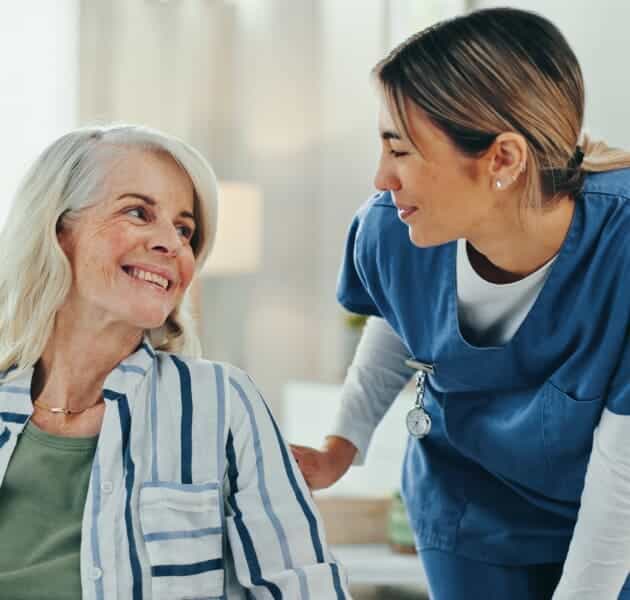 A nurse in blue scrubs smiles and leans toward an older woman with gray hair, who is sitting and smiling back at her. They appear to be having a friendly, supportive conversation in a bright room.