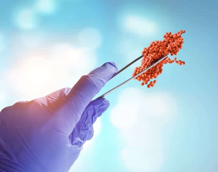 A gloved hand holds metal tweezers gripping a cluster of small red spheres, resembling a model of a virus or molecule, against a blue background with light spots.