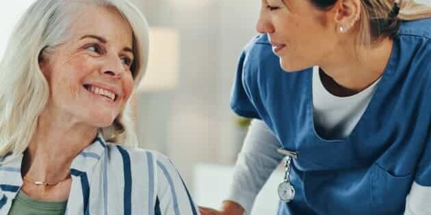 A nurse in blue scrubs smiles and leans toward an older woman with gray hair, who is sitting and smiling back at her. They appear to be having a friendly, supportive conversation in a bright room.