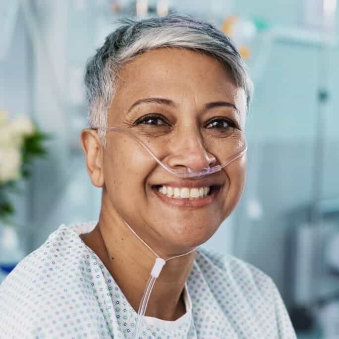 Smiling woman with short gray hair wearing a hospital gown and oxygen nasal cannula, seated in a medical setting with blurred medical equipment in the background.