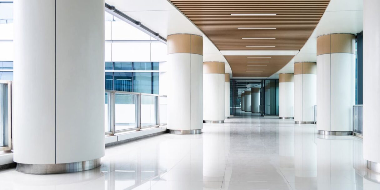 A modern, bright hallway with large white columns, glass railings, and a wooden slat ceiling. Sunlight streams in through tall windows, reflecting off the glossy white floor. The space is open and spacious.