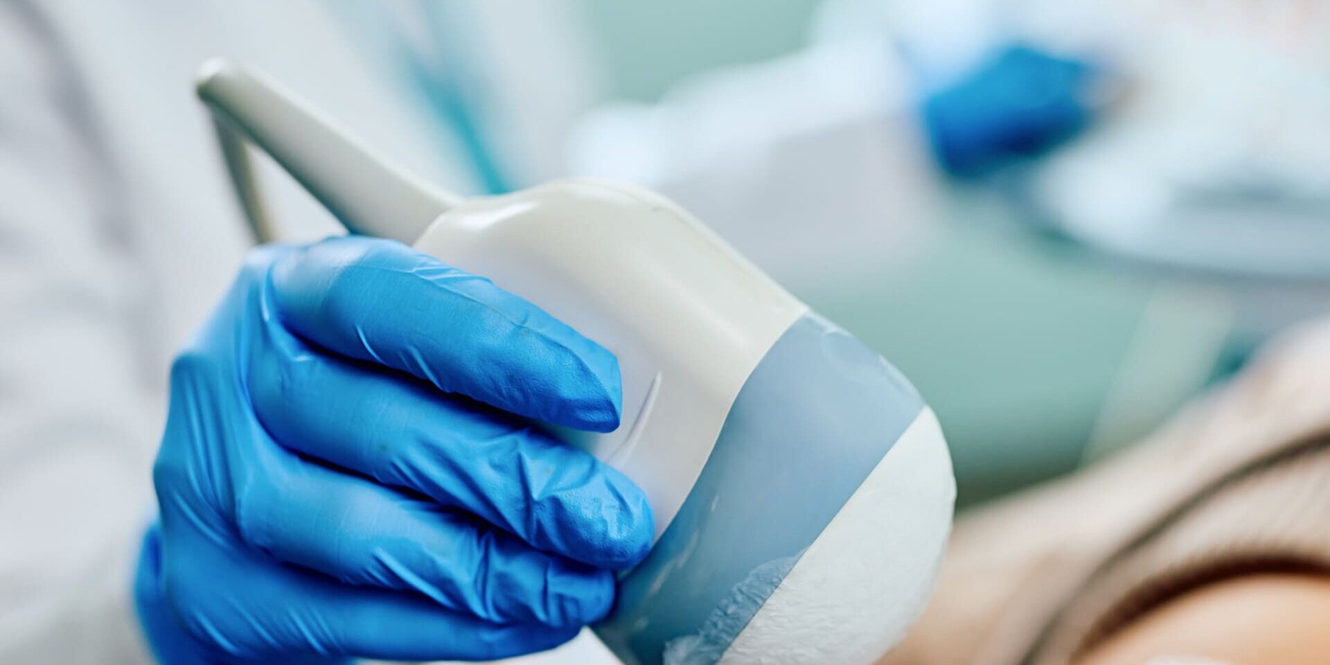 A medical professional wearing blue gloves holds an ultrasound probe, preparing to perform an ultrasound scan on a patients leg, which is covered in clear gel.