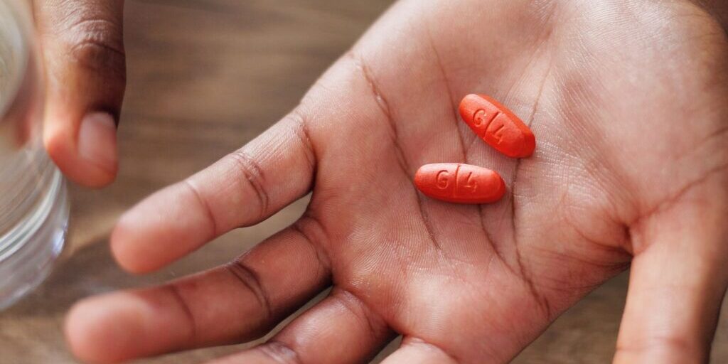 A person holds two oblong, orange pills marked G1 in their open palm, with a glass of water nearby on a wooden surface.