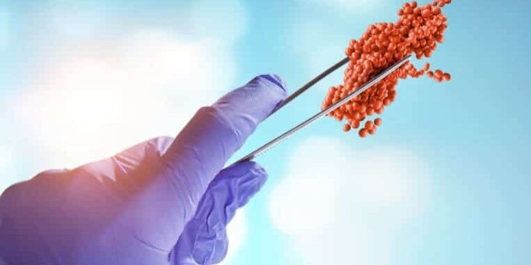 A gloved hand holds metal tweezers gripping a cluster of small red spheres, resembling a model of a virus or molecule, against a blue background with light spots.
