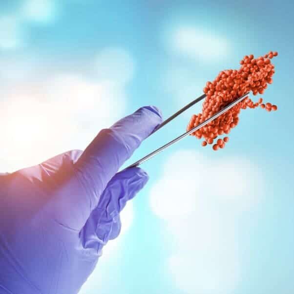 A gloved hand holds metal tweezers gripping a cluster of small red spheres, resembling a model of a virus or molecule, against a blue background with light spots.