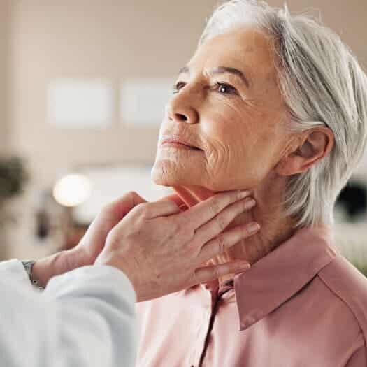 A doctor gently examines the neck of an older woman with short gray hair, who is wearing a pink blouse and looking slightly upward. The setting appears to be a medical office.
