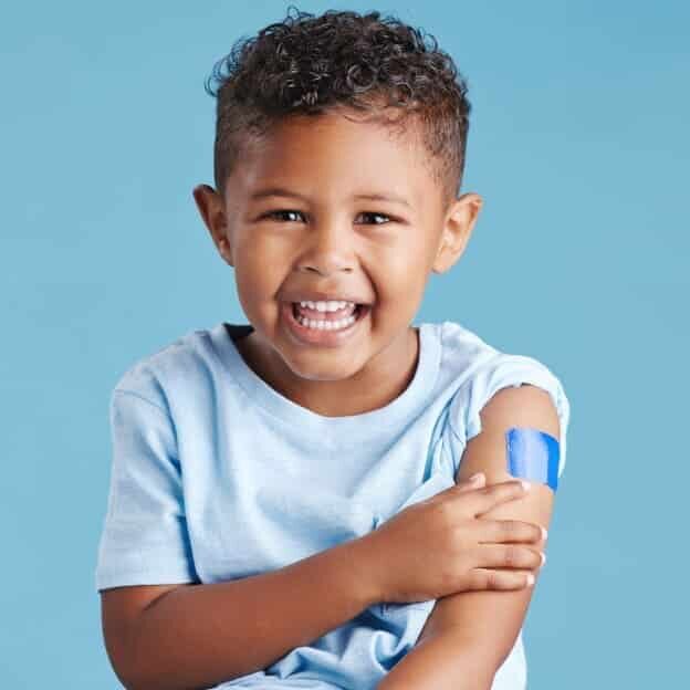 Smiling young boy with curly hair wearing a light blue shirt, holding his arm to show a blue adhesive bandage on his upper arm, set against a plain blue background.