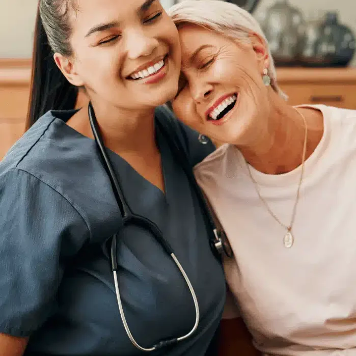 A smiling healthcare worker with a stethoscope sits closely with an older woman, both looking happy and relaxed, sharing a joyful moment together indoors.