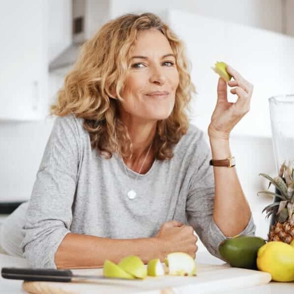 A smiling woman with wavy blonde hair sits in a bright kitchen, holding a piece of green apple. A cutting board with sliced apples, a pineapple, and a mango are in front of her.